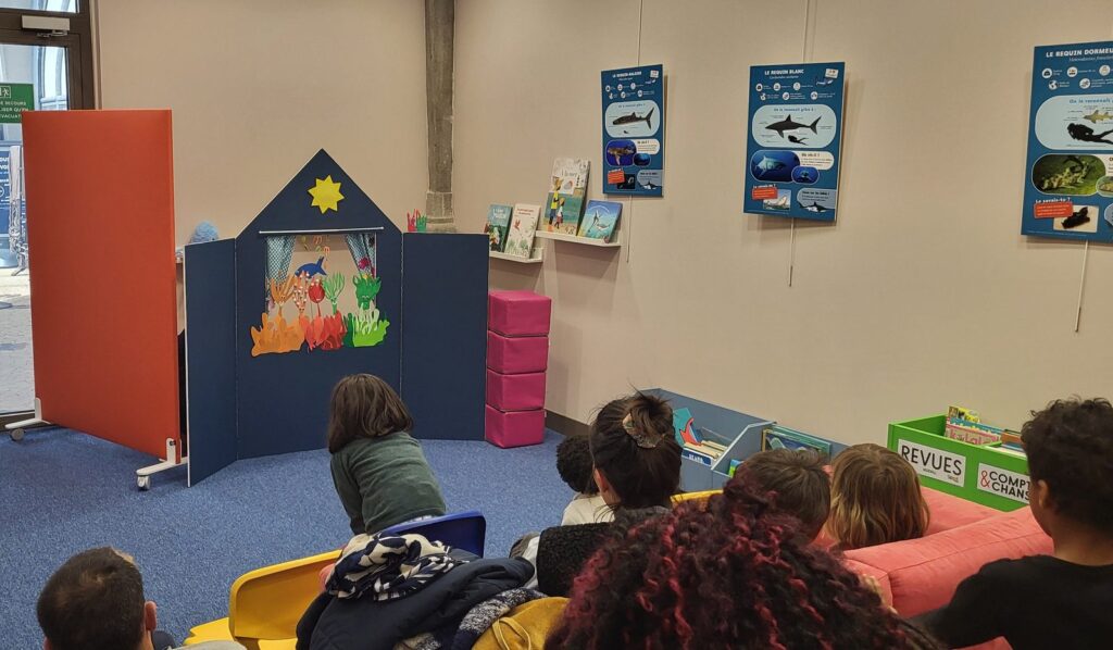 Des enfants assis de dos regardent un petit théâtre de marionnettes installé dans une médiathèque.