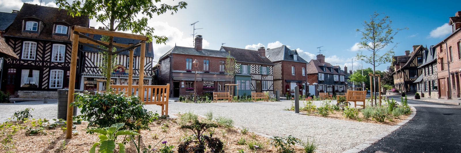 Place pavée d'un village normand bordée de maisons à colombages, avec des stands installés.
