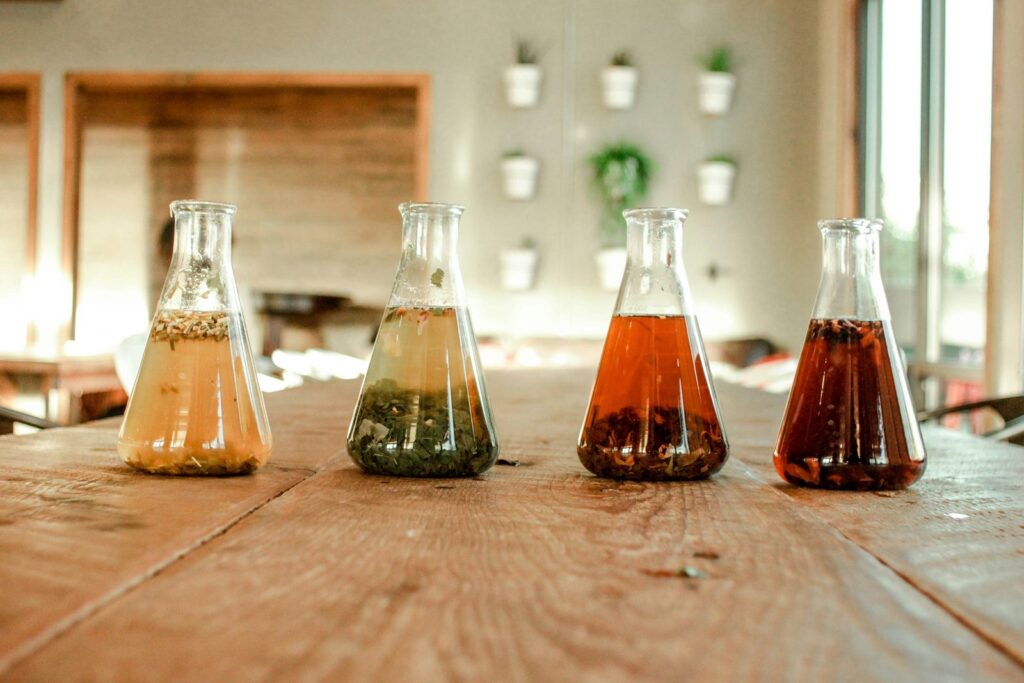 Quatre fioles de laboratoire remplies d'infusions de couleurs différentes, alignées sur une table en bois.