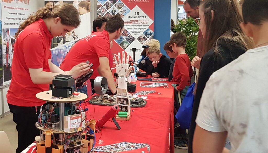Sur un stand, des étudiants en tee-shirt rouge font manipuler un bras robotisé à des visiteurs.