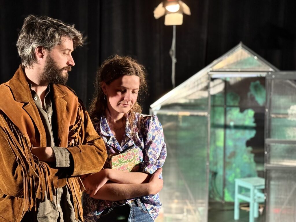 Deux comédiens se font face devant une serre en verre, sur un plateau de théâtre.