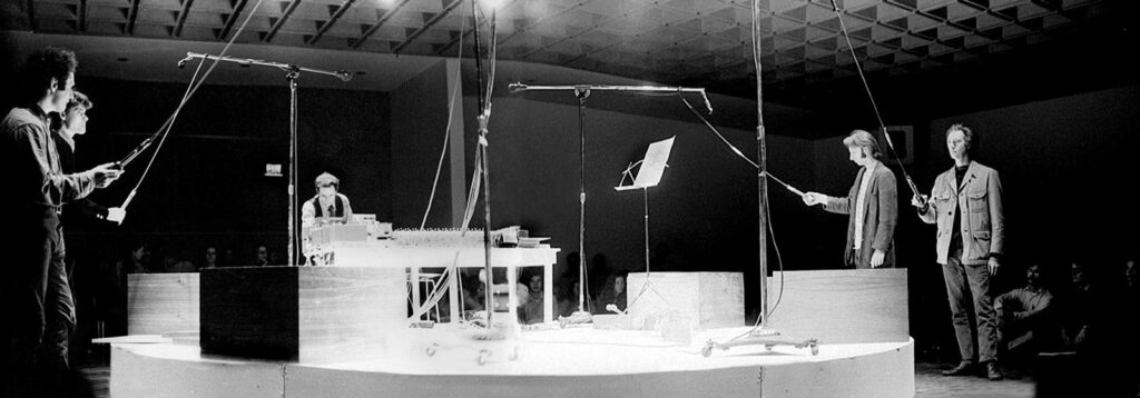 Photographie ancienne en noir et blanc d'une démonstration d'acoustique sur une scène, devant plusieurs personnes.