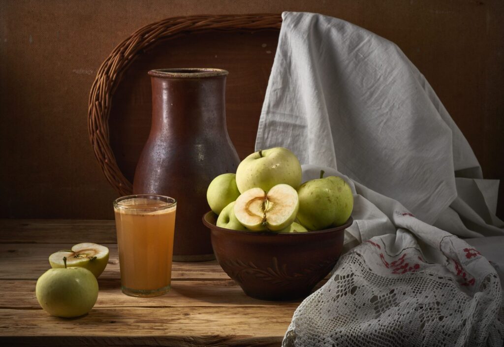 Pommes, verre de cidre et pichet de grès composés en nature morte sur une table en bois.