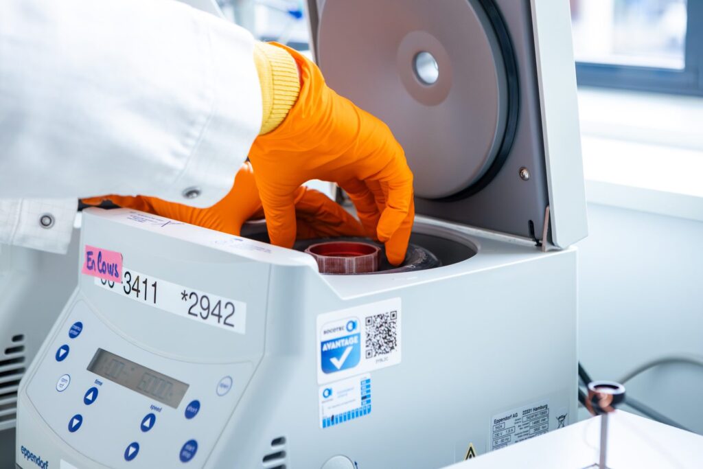 Une main gantée d'orange dépose un échantillon dans une centrifugeuse de laboratoire.