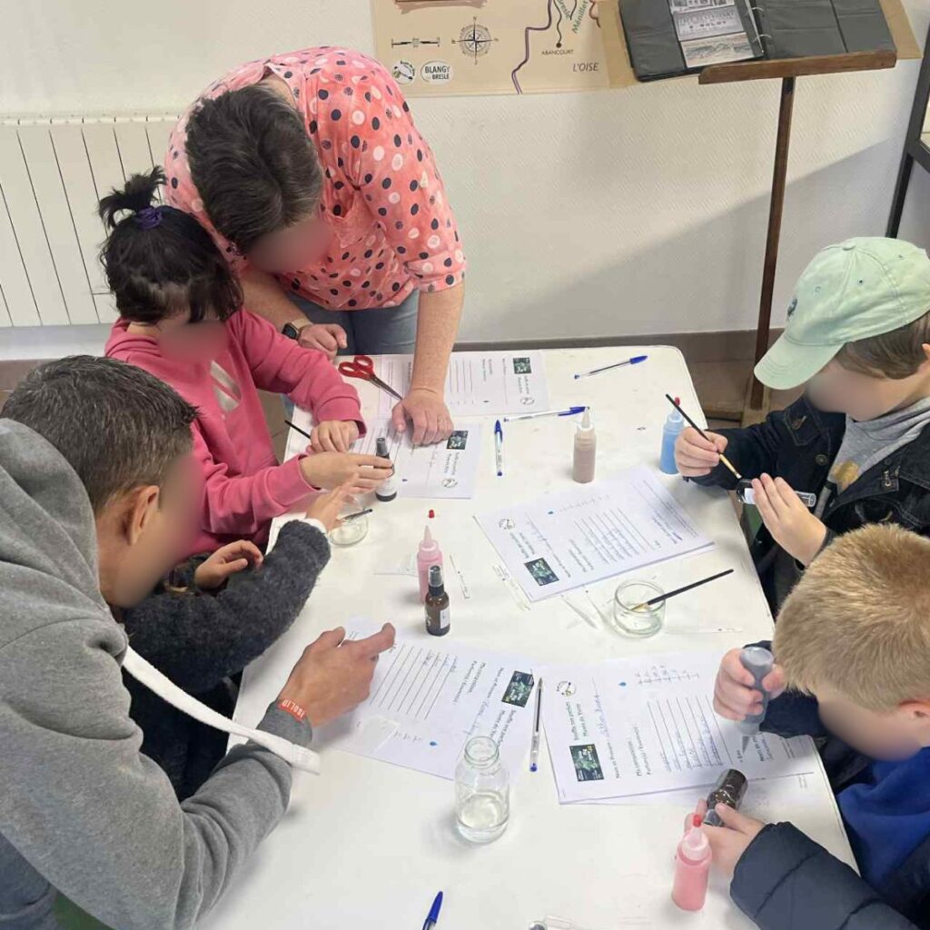 Des enfants manipulent des bandes de papier et des flacons autour d'une table d'atelier, accompagnés d'un adulte.