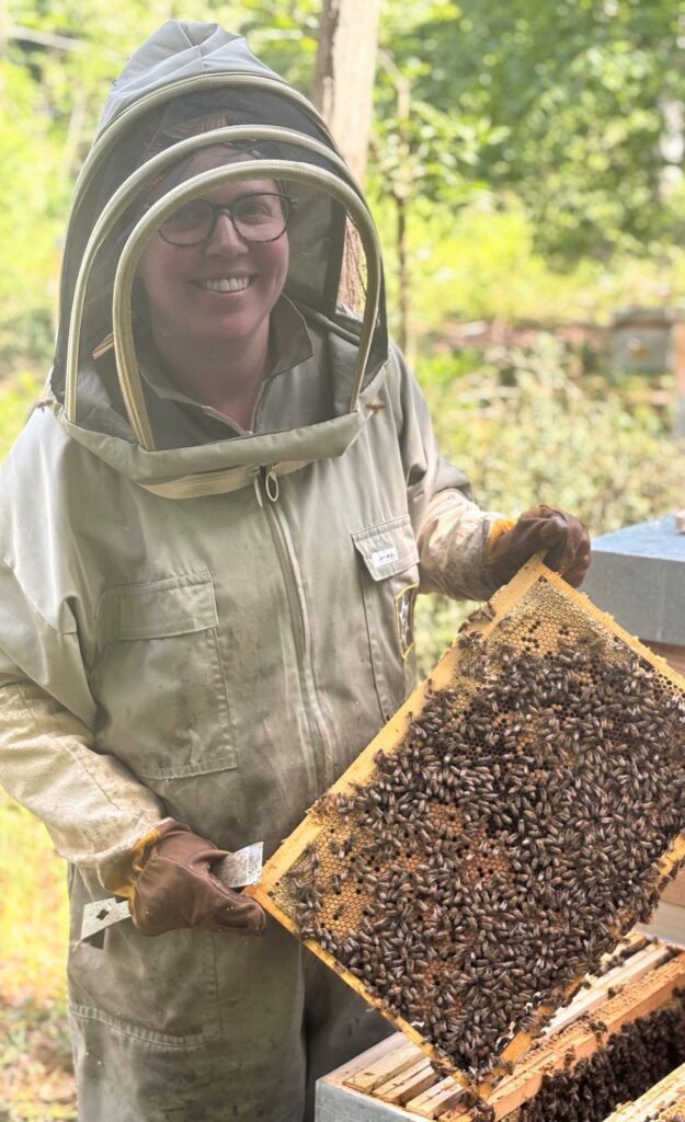 Une apicultrice en combinaison présente un cadre de ruche couvert d'abeilles.