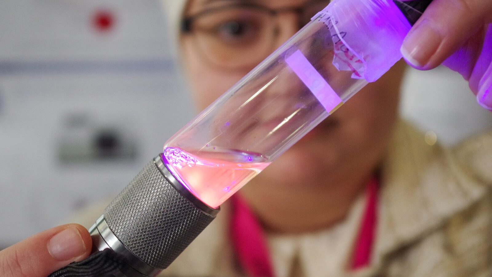 Une main tient une éprouvette dont le contenu est éclairé en violet par une lampe.