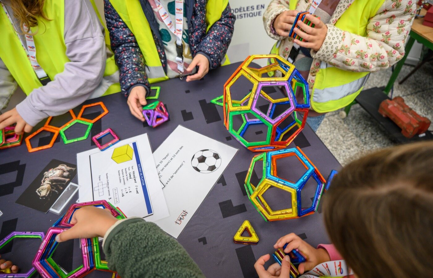 Des enfants assemblent des polyèdres avec des plaques magnétiques colorées autour d'une table d'atelier.
