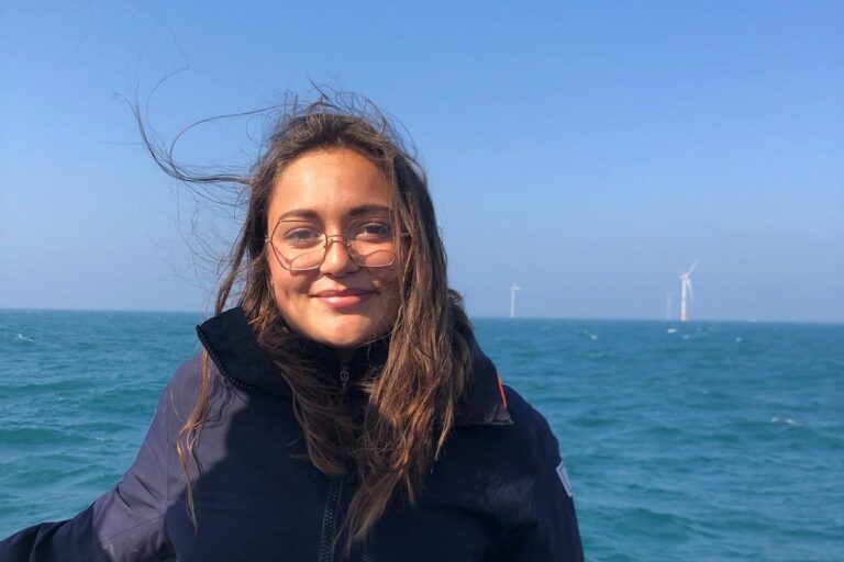 Mathilde Charbonnelle sur le pont d'un bateau, en mer, des éoliennes au loin.