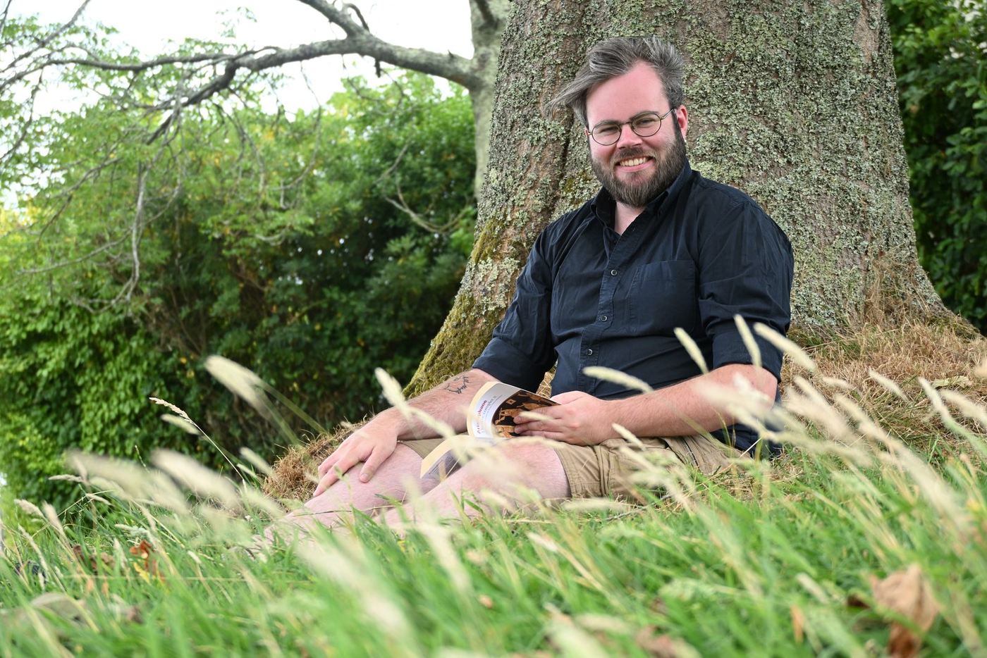 Raphaël Dubois, doctorant en histoire, assis dans l'herbe au pied d'un grand arbre.